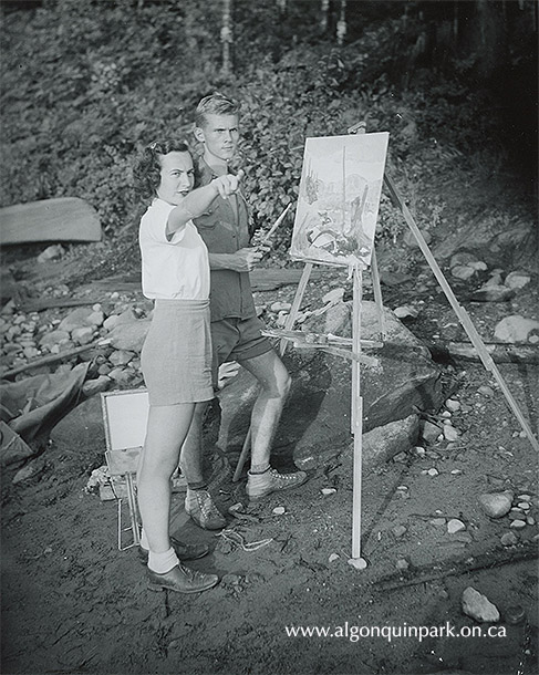Image: Barbara Caldwell and Robert Bateman, Algonquin Wildlife Research Station, 1949. Bateman stands holding a paint brush next to an easel with a landscape painting in progress. His supplies are visible on the ground behind them. APPAC, x2024.8.2.1. 