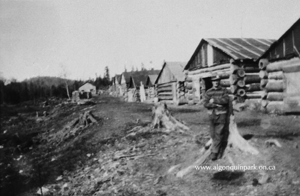Image: Man leaning against a stump in front of several buildings at the J.R. Booth Lumber camp at the north end of Shirley Lake, Algonquin Park, 1925. APPAC, 1976.74.3, Max Borutski. 